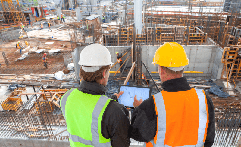 Zwei Bauarbeiter mit Schutzhelmen und Warnwesten betrachten ein Tablet auf einer Baustelle. Zwei Bauarbeiter mit Schutzhelmen und Warnwesten betrachten ein Tablet auf einer Baustelle.